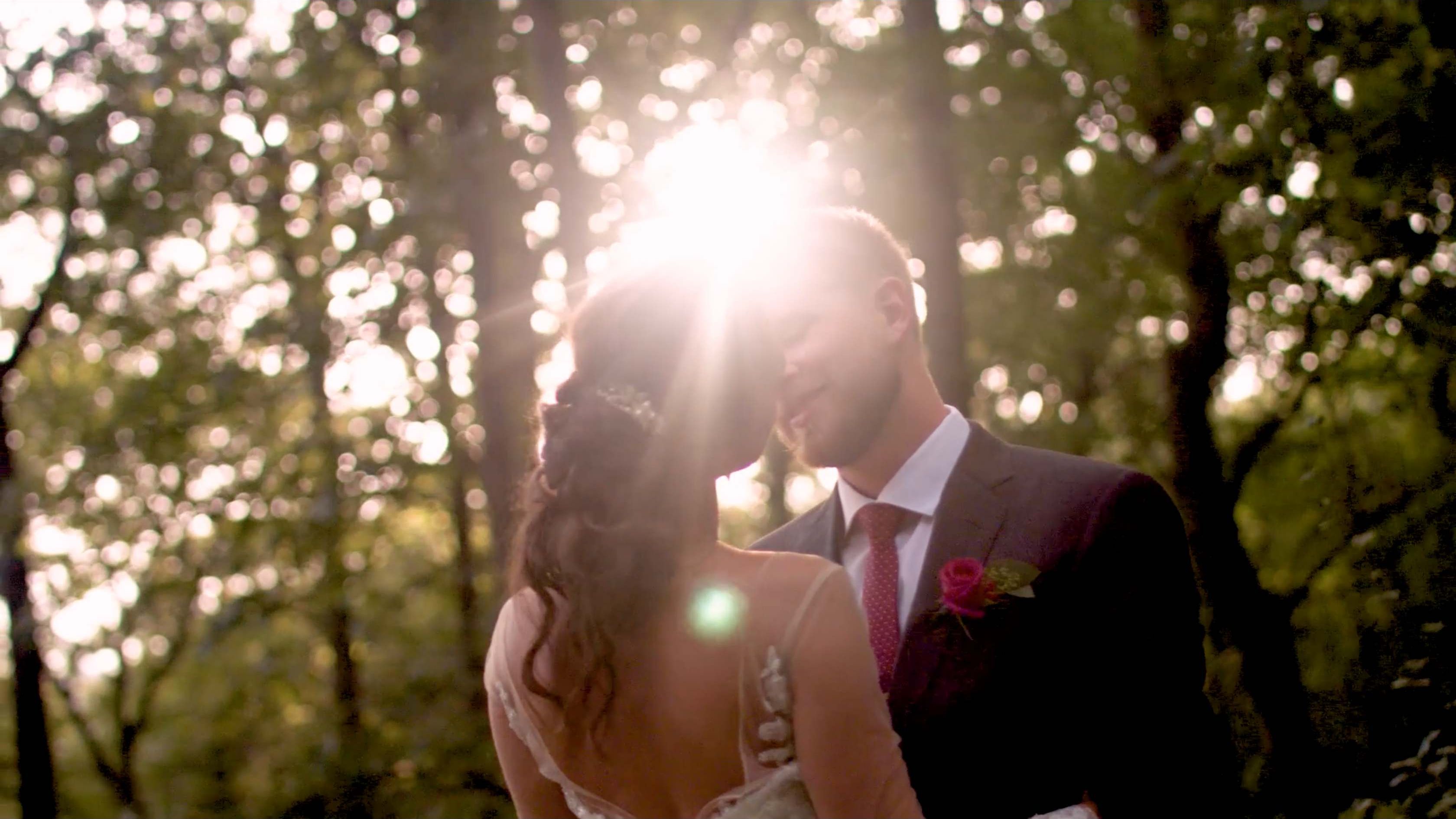 A bride and groom touching foreheads in a sunlit forest as light bursts between them